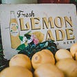 Fresh lemons sit in front of a  sign that says, “Fresh Lemonade sold here”.
