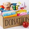 Box with Donation written across the front, filled with clothes, toys, and stuffed animals. Apple in front of box