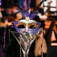 A purple Mardi Gras mask with green and gold sparkles and purple and black feathers sits in a martini glass with feathers and beads spilling out the sides of the glass.