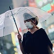 A Japanese woman wearing a medical mask and black blouse walks toward the left-center of the frame while carrying clear umbrella