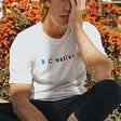 A young man is sitting on the ground with his hand pushed against the side of his face. He has the look of someone who lacks inspiration and creativity. And he’s wearing a white t-shirt that says B-CREATIVE written in rainbow colours.