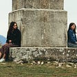Two women facing away from each other, sitting on a platform on either side of an ancient pillar.