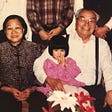 A young Jackie with a hilarious bowl cut in a pink dress, sitting with her maternal grandparents.