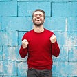 Man in red shirt cheers jubilantly in front of a blue brick wall