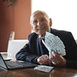 happy old man dressed in nice suit holding stack of money 100 dollar bills and sitting next to a computer