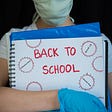 Child wearing a face mask and gloves, holding notebook with Back to School written on it. Photo by Amy Mitchell/Getty Images