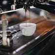 A shot of espresso, mid-pour dropping into a white ceramic cup under an espresso machine, (courtesy of unsplash)