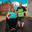 Photo by Maria Oswalt on Unsplash | A woman in a wheelchair and a man by her side holding a message “We are all humans”.