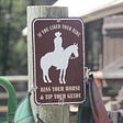 Sign on the fencepost of a corral, saying: If you liked your ride, kiss your horse and tip your guide. Pictured on the brown sign is the white silhouette of a horse and rider. Rider wears a cowboy hat.