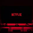 An image of the Netflix logo on a television screen in a dark room. The television is on top of a cabinet with a console gaming controller sitting in front of it. The red from the logo font resonates outward from the television giving a sort of ominous glow.