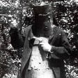 A black-and-white still showing an actor holding guns. His face is obscured by metal armor.
