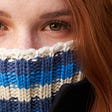 redheaded young woman has a turtleneck sweater pulled up over her mouth and nose, so that only her eyes are showing.