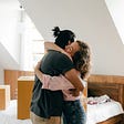 A young couple hugging each other with unpacked boxes on the bed behind them.