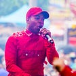 Todrick Hall, a young Black man, wearing a red shirt and ballcap, holding a microphone.