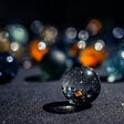 A group of marbles that are clear and in colors blue and orange sit atop a dark gray surface.