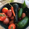 A green collander holding freshly-washed new potatoes, a little carrot, a small zucchini, two small cucumbers, and a handful of small tomatoes