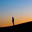 A shadowed figure of a man stands on a hill in front of an ombre background.