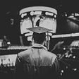 The black and white image shows the back of a masculine individual who is wearing a cap and gown, reminiscent of those worn for graduations in the US. The person overlooks a large stadium that is blurred into the background.