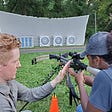 A young woman holds up a crossbow aimed at archery targets with the guidance of a man training her