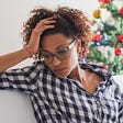A dark-skinned woman stares sadly at the ground. Her hand is on her head, just above her forehead. There is a Christmas tree behind her.