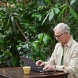 Elderly man sitting at a table with a laptop typing. There is a yellow cup behind the screen and green foliage around him.