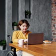 A woman sitting at her kitchen table, eating breakfast while working on her laptop