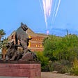 A metal statue featuring two wildcats in the foreground. Background: greenery, palm trees, university building, fireworks.