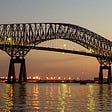 A photo of a large steel bridge over the immense Patapsco River in the early evening. The lights on buildings are visible in the distance. The river shows the reflections of the lights.