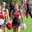 Three female teenage cheerleaders walk across a field carrying pompoms.