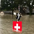 A flooded suburban American street; a partially submerged Swiss flag sits in the foreground; atop the flag dances a version of Monopoly’s Rich Uncle Pennybags, who has removed his face to reveal a grinning skull. Image: Ready.gov (modified) https://www.ready.gov/floods