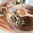 the image is of a wooden bowl and spoon in which are dried sage leaves; next to the bowl is a bunch of twig/roots knotted with a vine. The root is kakangda. The scene is very country, rustic, mystical.