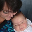 A white woman with dark hair rests her cheek on the head of her sleeping infant