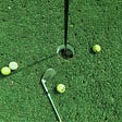 Golf balls scattered around the hole, with a club laying next to them.