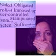A sad woman with bruises on her face and her hand over her mouth and over her head a poster with the words manipulated, over-controlled, restricted.