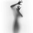 A photograph of an individual, with their hand pressed against the surface, is struggling to break free from the foggy white glass. Most of the photo is just a white background with just the trapped individual in the center. The individual’s face is unseen and the dark silhouette of the rest of their body can only be seen in the photo.