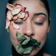 A women with her eyes closed who has leaves covering her mouth and wilted flower petals on the side of head