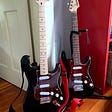 Squier Affinity Stratocaster on the left; black with maple neck, tortoise shell pickguard, white pickup covers and knobs. Mini-Squier on the right; black body with Pau Ferro neck, white pickup covers and knobs.