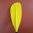A bright yellow boat paddle leaning against a red wall