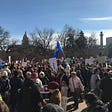 Crowd of people outside holding signs with a capital building in the background under the blue sky.