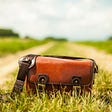 A red leather satchel sitting in the grass between two dirt roads.