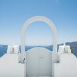 The image shows a gate with an arc in Oia, Greece. In the background you will find blue sky and a view of the sea.