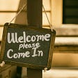 A chalk welcome sign bordered by rustic wood and hung with rope says Welcome, Please Come In