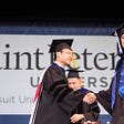 A broadly smiling 2022 graduate receives her diploma from the chair of her department at St. Peter’s University commencement ceremony. A large banner with the university’s name hangs in the background.