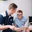 Ned (young male) from the Blitzit Team is showing a participant papers on how Blitzit can help with their NDIS plan. Ned is at the front in a Blue & White Blitzit shirt and the Male Participant who wears glasses and has red hair, wearing a light blue patterned shirt, is looking at the papers. They sit on a plain background.