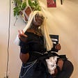 Ebony M. poses for a picture in a cosplay of Misa Amane from Death Note. They are wearing a blonde wig & a black goth dress.