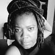 the image is a black and white selfie of the author of this piece, Ilis Trudie Palmer. She is staring into the camera with large expressive eyes. She wears dreadlocks that are wrapped on the top of her head resembling a turban.