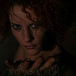 Woman with dark eyeliner starting menacingly at the camera with an upturned hand outstretched, against a dark background.