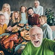 A multi-generational family smiles while posing for a group selfie around the table filled with their holiday feast.