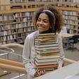 A young woman holding a pile of books in a library looks happy