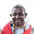 A picture of a Black, blind person with short hair. They are wearing necklaces in tones of yellow, red black and white, and a pink scarf around their shoulders. Photo credit: Canva